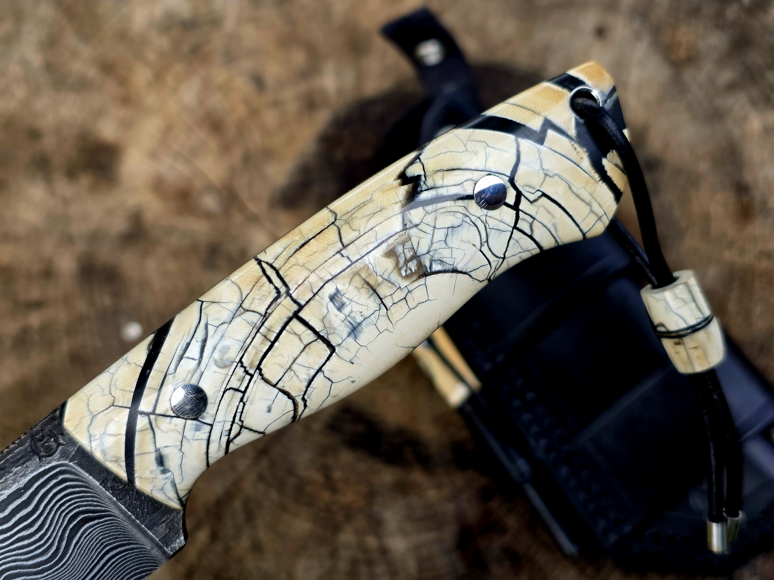 Bady K – Bushcraftmesser mit Mammutelfenbein-Griff Bushcraftmesser Bady K mit Griff aus stabilisiertem Mammutelfenbein, schwarzer Maserung, Damastklinge und Fangriemen.