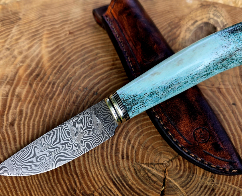 Panah – Balbach-Damast mit blauem Hirschhorn Handgefertigtes Panah Jagdmesser aus Balbach-Damast mit blau stabilisiertem Hirschhorngriff, Neusilber und Vulkanfiber, auf brauner Lederscheide.