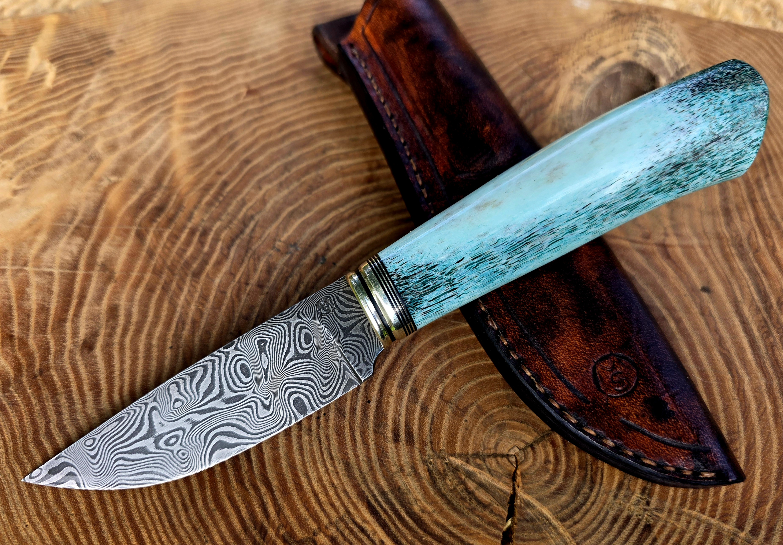 Panah – Balbach-Damast mit blauem Hirschhorn Handgefertigtes Panah Jagdmesser aus Balbach-Damast mit blau stabilisiertem Hirschhorngriff, Neusilber und Vulkanfiber, auf brauner Lederscheide.