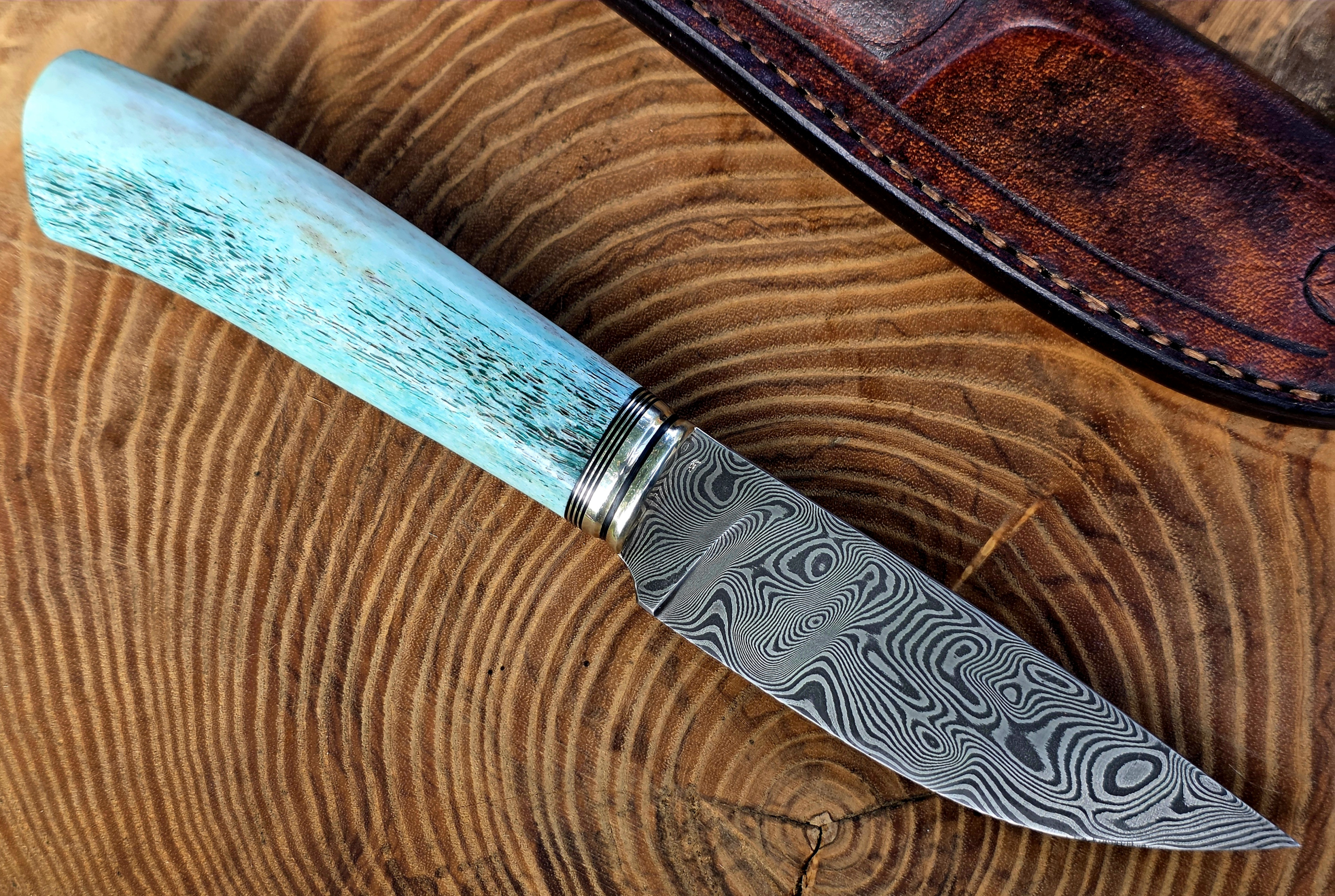 Panah – Balbachdamast und blaues Hirschhorn Handgefertigtes Panah Jagdmesser mit Balbachdamast-Klinge, blau stabilisiertem Hirschhorngriff und brauner Lederscheide.